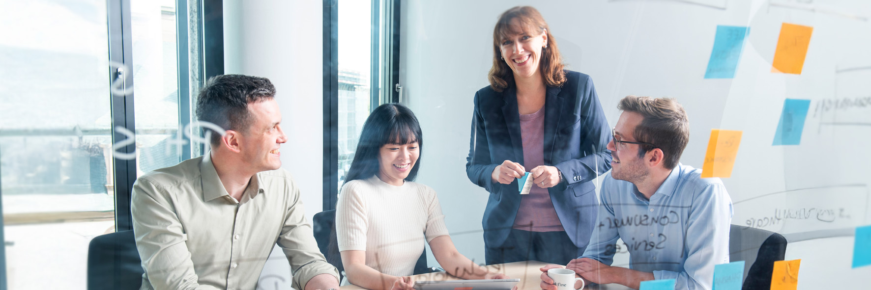 Four people in business attire are in a modern office in front of a glass wall covered with sticky notes and markings. Three of them are seated at a table, engaged in conversation, while the fourth person stands behind the table, smiling at the camera.