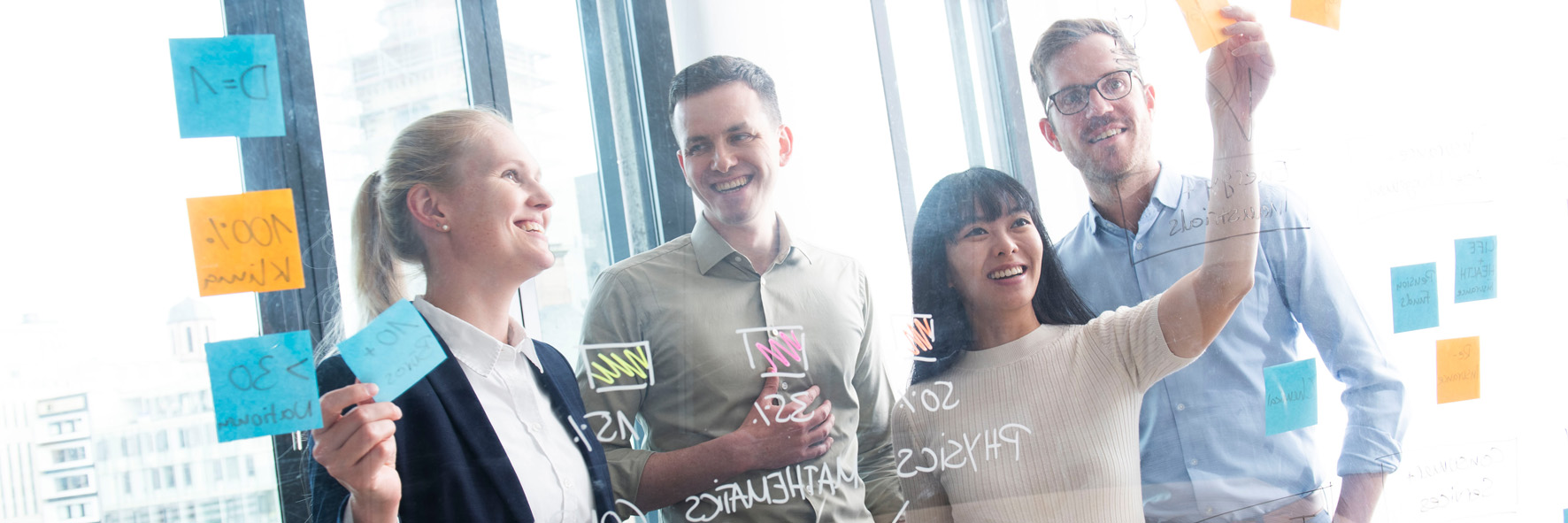 Vier Personen in Businesskleidung stehen in einem modernen, hellen Büro vor einer Glaswand, die mit bunten Haftnotizen und Markierungen versehen ist. Alle wirken konzentriert und arbeiten gemeinsam an einer Aufgabe.
